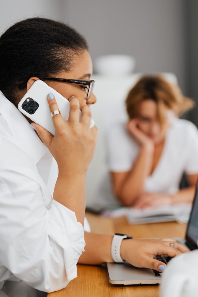 Black woman using cellphone and laptop at work, multitasking indoors.